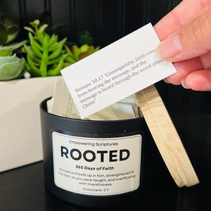 May include: A black container labeled "ROOTED" with a wooden lid, holding small cards. The cards have text from Romans 10:17. The container is on a black surface, with greenery in the background.