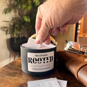 May include: A black container labeled "Man of Faith" with the word "ROOTED" in large letters. The container holds small white cards, and a hand is reaching in to select one. The text "Empowering Scriptures" is also visible.
