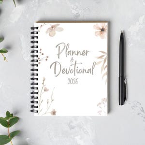 May include: A white spiral-bound planner and devotional for 2026 with floral accents and the text "Planner & Devotional". A black pen rests beside the planner on a gray surface.