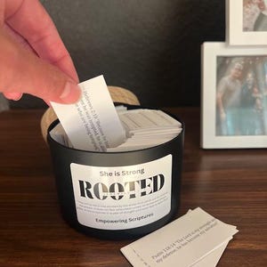 May include: A black container labeled "She is Strong ROOTED Empowering Scriptures" filled with small white cards. A hand is pulling a card from the container. The cards have text on them. The container sits on a wooden surface.