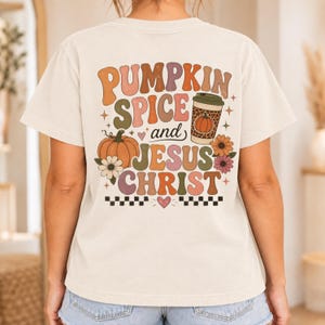 May include: A cream-colored t-shirt featuring a back graphic with the text PUMPKIN SPICE and JESUS CHRIST in retro-style lettering. The design includes a pumpkin, a leopard-print coffee cup, flowers, small hearts, stars, and a checkered border.