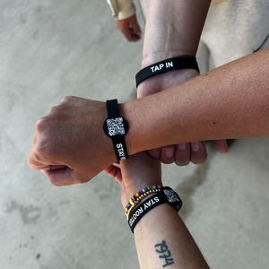 May include: Several black wristbands are displayed, each with white text. One reads "TAP IN," while others feature QR codes and the words "STAY ROOTED." A beaded bracelet with colorful beads is also visible. The image is taken from a slightly elevated angle.