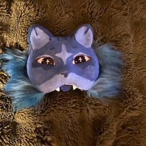 May include: A blue cat mask with a textured surface, featuring pointed ears, cross-shaped pupils, and a snarling mouth with white teeth. The mask is surrounded by blue faux fur.