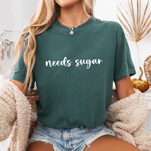 May include: A teal t-shirt with the words "needs sugar" in white script. The short-sleeved, crew neck shirt is worn with denim shorts and a beige cardigan. The image has a casual, relaxed feel.
