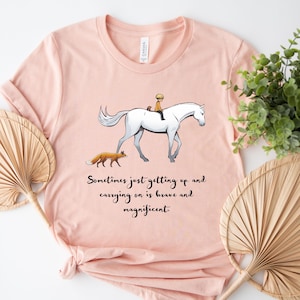 May include: Pale pink t-shirt with an illustration of a white horse, rider, and fox. The text says, "Sometimes just getting up and carrying on is brave and magnificent."