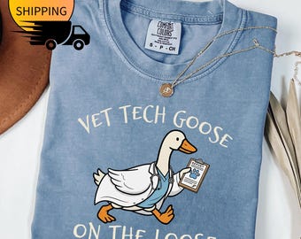 May include: A light blue t-shirt with the text "VET TECH GOOSE ON THE LOOSE" and a cartoon goose wearing a lab coat and holding a clipboard. The shirt has a crew neck and short sleeves. A gold necklace is draped across the shirt.