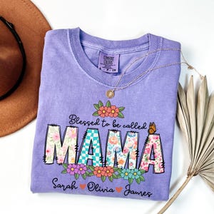 May include: A lilac Comfort Colors t-shirt with the text "Blessed to be called MAMA" in a floral design. The shirt includes the names Sarah, Olivia, and James. A gold necklace and a brown hat are also visible.