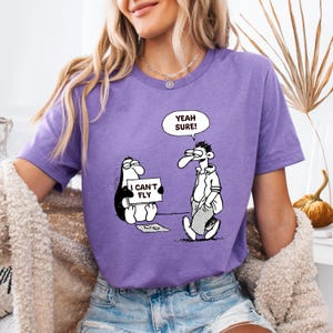 May include: A purple t-shirt featuring a cartoon illustration. The graphic shows a penguin holding a sign that reads "I Can't Fly" and a man with a speech bubble saying "Yeah Sure!"