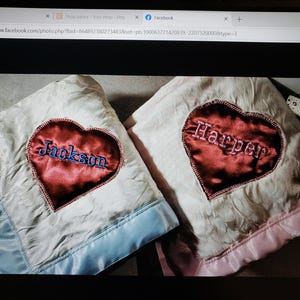 May include: Two soft blankets with a textured white surface and satin trim. One blanket features a dark red satin heart patch embroidered with the name Jackson in blue thread. The other has a matching heart patch with the name Harper in pink.