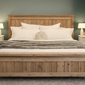 May include: A wooden bed frame with a headboard and footboard. The bed is made of light brown wood and has a plush, gray blanket. White pillows and a decorative round pillow are on the bed. Two bedside tables with lamps are on either side.