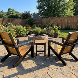 May include: Two wooden outdoor chairs and a small side table. The chairs have a black frame and natural wood seat and back. The set is on a stone patio with a fire pit and garden in the background.