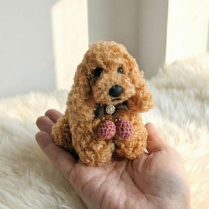 Puede incluir: Una figura de perro en miniatura hecha a mano, con pelo rizado marrón. El perro tiene una nariz negra y lleva un collar con dos pompones rosas. La figura está colocada en una mano.