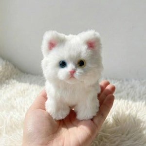 May include: A small, fluffy white cat figurine with one blue and one yellow eye. The cat has pink accents on its ears and nose. The figurine is held in a person's hand, and the background is a soft, white, textured surface.