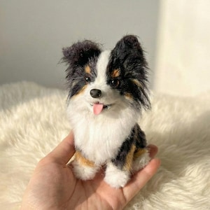 May include: A small, plush dog figurine with a black, white, and brown coat. The dog has a pink tongue sticking out and is sitting on a fluffy white surface. The figurine is held in a person's hand.