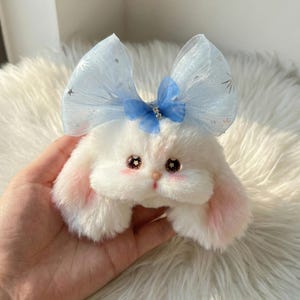 Könnte beinhalten: Ein weißes Plüschkaninchen mit rosa Innenohren und rosigen Wangen. Es hat große, funkelnde Augen und eine blaue Schleife mit einer kleineren Schleife und einem Schmuckstein in der Mitte. Das Kaninchen wird in einer Hand vor einem flauschigen weißen Hintergrund gehalten.