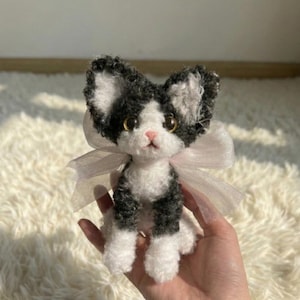 May include: A handmade plush cat toy with black and white fur, gold eyes, and a pink nose. The cat has a light pink ribbon tied around its neck. The toy is held in a person's hand.