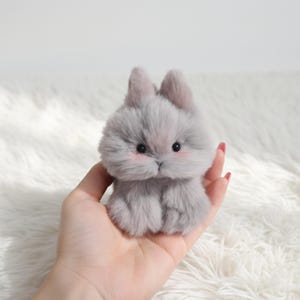May include: A small, plush gray bunny with pink cheeks and dark eyes. The bunny has upright ears and a fluffy body. The toy is designed to look like a real rabbit. The bunny is a cute, handmade item.