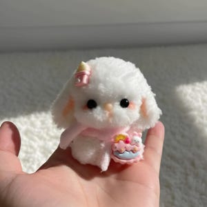 Könnte beinhalten: Ein kleines, weißes Plüschtier in Form eines Hasen mit schwarzen Augen und rosigen Wangen. Es trägt einen rosa Schal und eine rosa Schleife. Der Hase hält einen Miniaturkuchen mit bunter Glasur und einem kleinen Bären. Das Spielzeug wird in einer Hand gehalten.