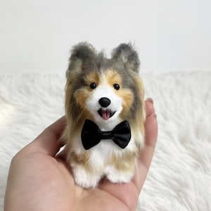 May include: A miniature Shetland Sheepdog toy, complete with a black bow tie. The dog's fur is a mix of brown, white, and grey. Its mouth is open, showing a pink tongue. The toy is held in a person's hand.