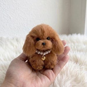 Op de afbeelding: Een miniatuur bruin poedel pluche speelgoed met een parelketting en een gouden hartvormige hanger. Het speelgoed heeft een zachte textuur en donkere ogen. De poedel zit op een witte, zachte ondergrond.