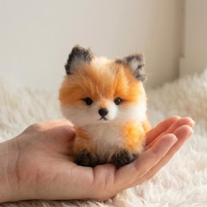Op de afbeelding: Een kleine, zachte vossenknuffel met oranje, witte en zwarte vacht. De vos wordt in een open hand gehouden, wat de gedetailleerde kenmerken en zachte textuur benadrukt. Het speelgoed heeft een schattig, levensecht uiterlijk.