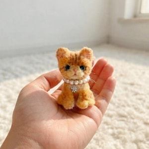 Pode incluir: Uma pequena figura de gatinho malhado laranja feito de feltro de agulha, com pelo texturizado, olhos escuros e nariz rosa. O gatinho usa um colar de pérolas brancas falsas com um pequeno pingente prateado em forma de flor no centro.