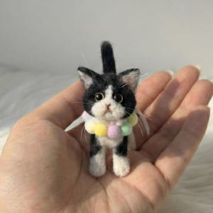 May include: A miniature felt cat, predominantly black and white, adorned with a multi-coloured pom-pom collar. The cat has large eyes and a fluffy texture. This handcrafted figurine highlights the detail and skill involved in creating handmade items.