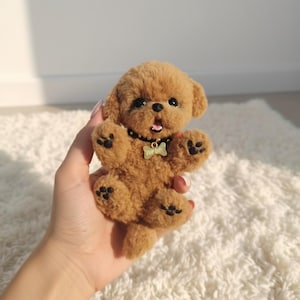 以下が含まれることがあります： 黒い足跡と「TOP DOG」と書かれた骨型のタグが付いた黒い首輪が付いた茶色のぬいぐるみ犬。おもちゃの犬は口を開けており、足を上げた状態で配置されています。おもちゃは白いふわふわの表面にあります。
