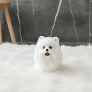 Op de afbeelding: Een witte Pomeranian hondenfiguur met zwarte ogen en een roze tong. De hond zit op een wit, getextureerd tapijt. De vacht van de hond is lang en zacht en zijn oren zijn roze.
