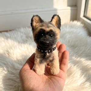 May include: A small, needle-felted dog figurine with tan and black fur, pointed ears, and a dark muzzle. The dog wears a black collar featuring small silver studs and a tiny, dangling silver bell, crafted with a textured, fuzzy appearance.
