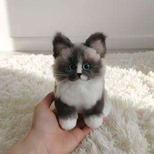 Op de afbeelding: Een zittend pluche kattenbeeldje met grijs en wit haar, blauwe ogen en lange witte snorharen. De kat heeft grijze oren en poten, en een witte borst en buik. Het beeldje wordt in een hand gehouden.