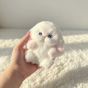 Könnte beinhalten: Ein flauschiges, weißes Stoffkaninchen mit blauen Augen und rosa Akzenten an Ohren und Pfoten. Das Plüschtier hat einen runden Körper und ist als Babykaninchen gestaltet. Ein niedliches und kuscheliges Spielzeug.