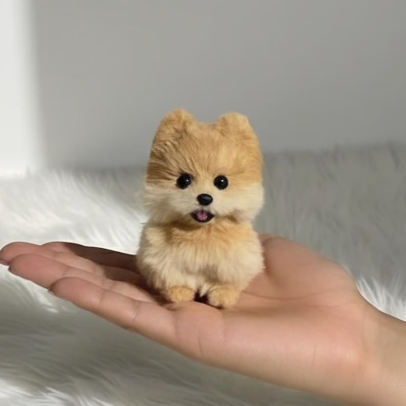 Op de afbeelding: Een kleine, pluizige Pomeranian-hondfiguur met bruin en cr&egrave;mekleurig haar. De hond heeft zwarte ogen en een kleine roze tong die uitsteekt. De figuur wordt in de hand van een persoon gehouden.