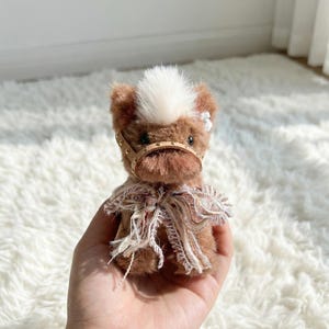 以下が含まれることがあります： 白い前髪と革のハーネスが付いた、小さな茶色のぬいぐるみ馬。 馬の首には装飾的なスカーフが付いています。 おもちゃは人の手で持たれています。