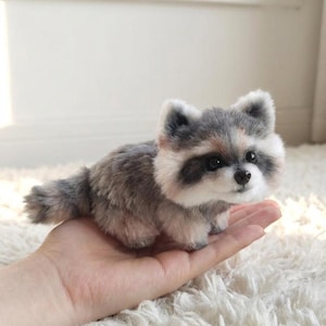 May include: A small, handcrafted raccoon figurine made of soft, textured grey and white faux fur. The creature features a black nose, dark eyes, and the characteristic black mask markings around its eyes, with a fluffy tail and pointed ears.