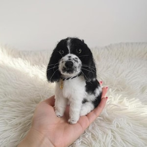 May include: A miniature dog figurine, hand-crafted with black and white detailing. The dog features long, floppy ears, a black nose, and a small gold-coloured tag on its collar. The figurine is held in a hand against a fluffy white background.