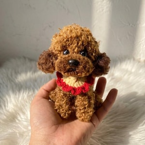 May include: A small, brown, curly-haired crocheted poodle figurine featuring a textured, yarn-based coat, dark plastic eyes, a black nose, and a decorative collar made of a cream-coloured centre with a ruffled, bright red crocheted border.