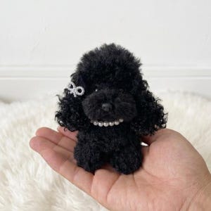 Puede incluir: Una pequeña figura de caniche negro con pelo rizado, un collar de perlas y un lazo decorativo. El juguete se sostiene en una mano, mostrando su tamaño y detalle. El fondo es una superficie blanca y suave.