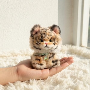 May include: A small, fluffy plush tiger cub with orange, black, and white fur. The cub has large dark eyes, a small pink nose, and pointed ears. It wears a light green collar featuring a small, dangling green leaf-shaped charm.