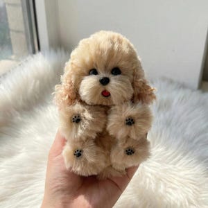 May include: A plush toy dog with a light brown, fluffy coat and black paw prints on its paws. The dog has black eyes and a red tongue. The toy is designed to resemble a poodle or similar breed.