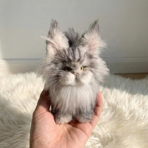 May include: A small, fluffy cat figurine in shades of grey and white, with detailed fur. The cat has upright ears and a slightly grumpy expression. The figurine is held in a hand, showing its small size and realistic appearance. The cat's fur has a soft, textured appearance.