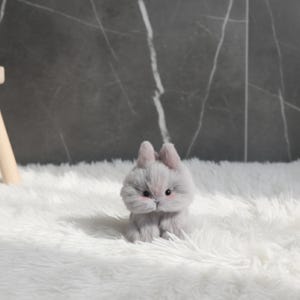 May include: A small, plush gray bunny with pink accents on its cheeks and ears sits on a fluffy white surface. The bunny has black eyes and a slightly furrowed brow, giving it a curious expression. The background is a dark gray marble-like wall.