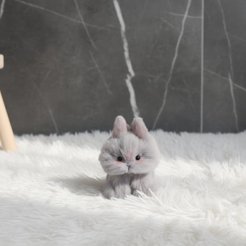 May include: A small, plush gray bunny with pink accents on its cheeks and ears sits on a fluffy white surface. The bunny has black eyes and a slightly furrowed brow, giving it a curious expression. The background is a dark gray marble-like wall.