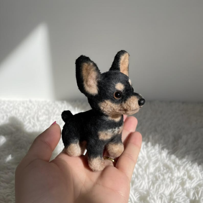 May include: A handcrafted miniature dog figurine. The small dog has black and tan fur, with large, upright ears. The figurine is held in a hand, showcasing its size and detail. The dog's eyes are dark, and its nose is black.