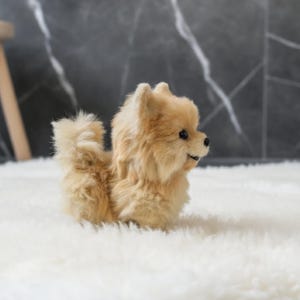 Op de afbeelding: Een pluche, bruin gekleurd Pomeranian hondenspeeltje met een pluizige staart en vacht. Het speelgoed heeft zwarte ogen en een zwarte neus en zit op een witte, zachte ondergrond. De hond kijkt naar rechts.