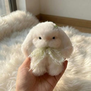 Könnte beinhalten: Ein kleines, flauschiges, weißes Stoffkaninchen mit schwarzen Augen und einer hellgrünen Schleife. Das Kaninchen hat lange, schlaffe Ohren und einen kleinen grünen Akzent auf dem Kopf. Das Plüschtier wird in einer Hand gehalten.