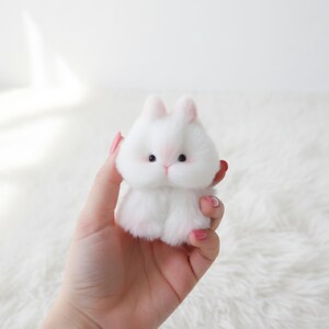 May include: A small, fluffy white bunny plush toy with pink accents on the ears, nose, and paws. The bunny has black eyes and a round, cuddly shape. The toy is held in a hand against a white background.