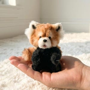 May include: A small, fuzzy red panda figurine with a white face, black nose, and black chest. The creature has pointed ears with white fur accents and a soft, textured appearance. It is crafted with a mix of reddish-brown and black fur tones.