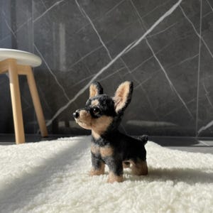 May include: A handcrafted miniature dog figurine, featuring a black and tan color scheme. The small dog is standing on a white, fluffy surface. The figurine has detailed features, including upright ears and a small tail.