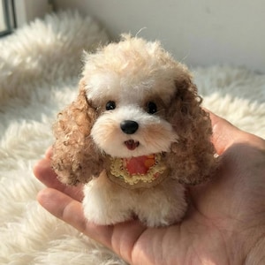 May include: A small, plush toy poodle with curly brown and cream-colored fur. The toy has a black nose, dark eyes, and a small red and yellow collar. The toy is held in a person's hand.